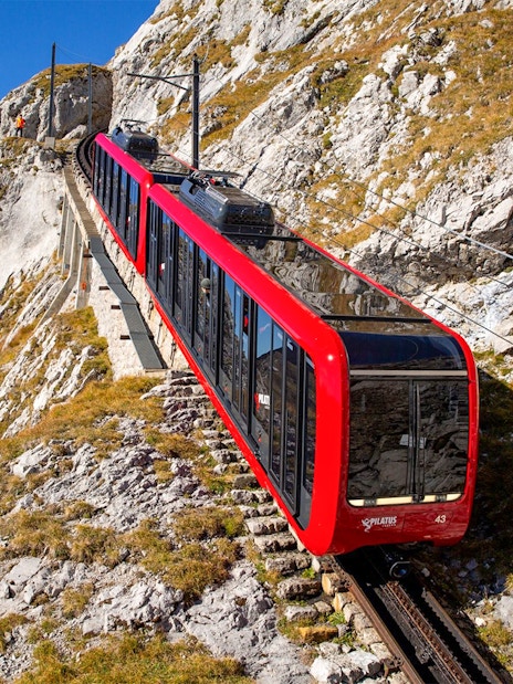 Red cogwheel train ascending Mount Pilatus, Switzerland, with rocky landscape.