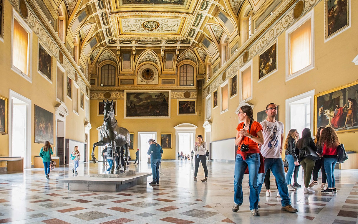 Visitors exploring the grand hall of the Archaeological Museum of Naples with statues and paintings.