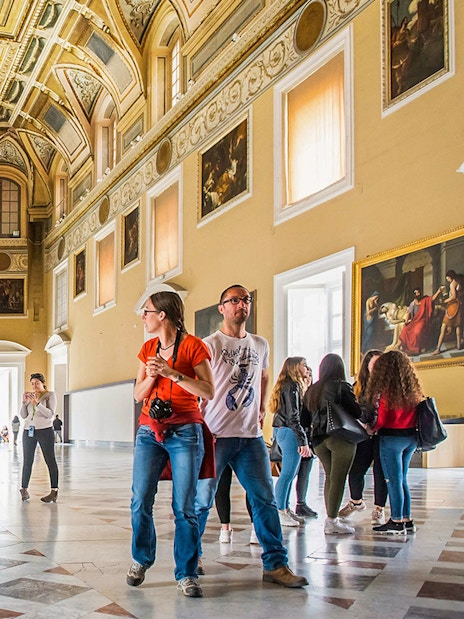 Visitors exploring the grand hall of the Archaeological Museum of Naples with statues and paintings.