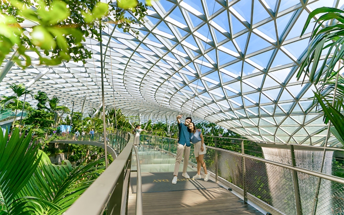 Couple taking selfie on walkway at Jewel Changi Airport, Singapore, under glass dome.