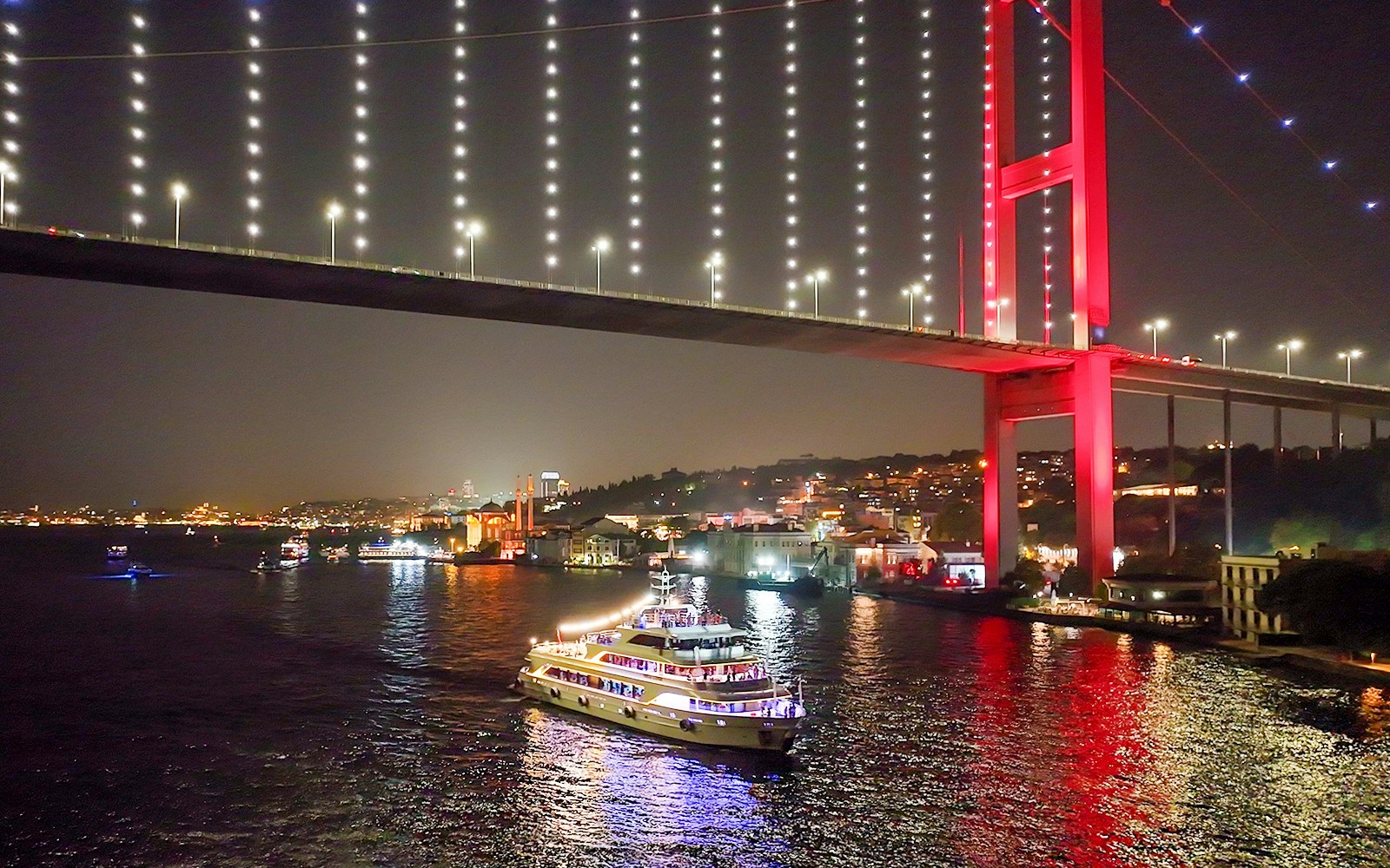 Bosphorus dinner cruise ship under illuminated bridge at night in Istanbul.