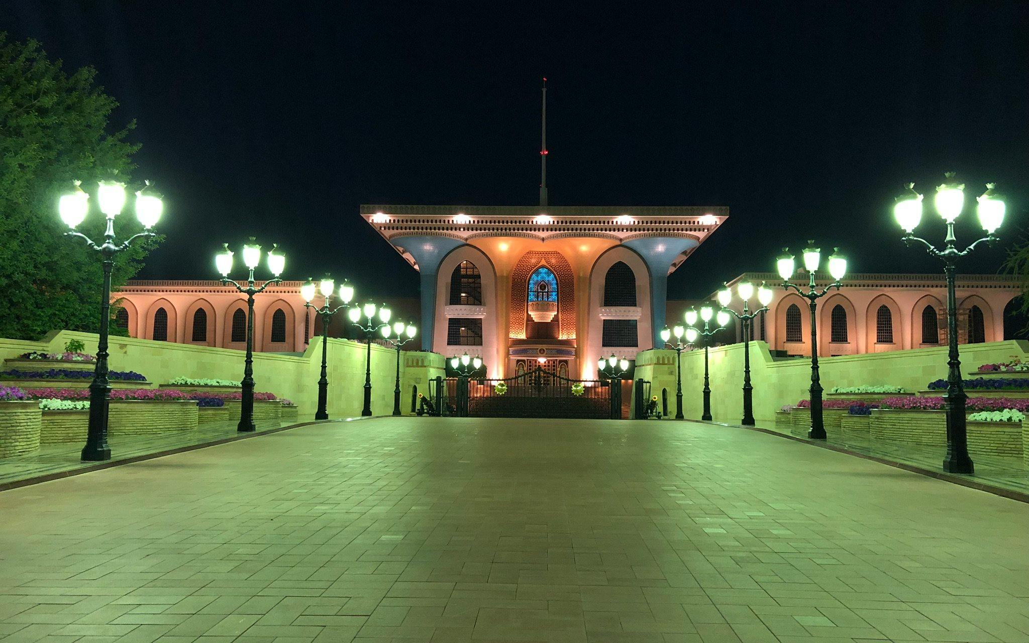 Al Alam Palace illuminated at night in Muscat, Oman, with surrounding gardens and lights.