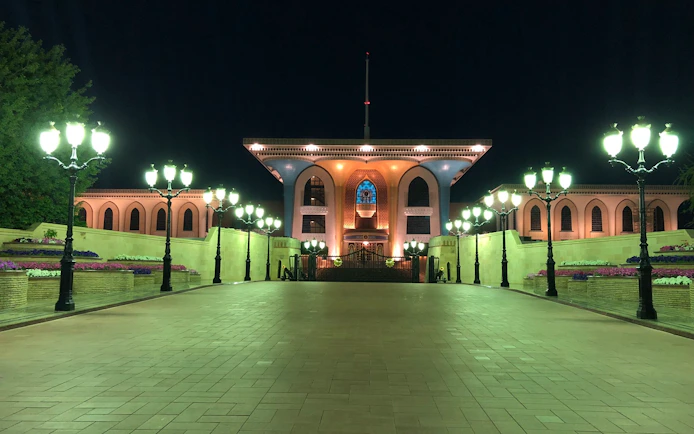 Al Alam Palace illuminated at night in Muscat, Oman, with surrounding gardens and lights.