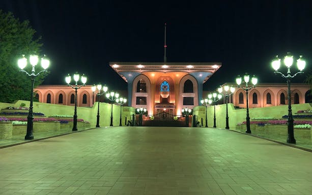 Al Alam Palace illuminated at night in Muscat, Oman, with surrounding gardens and lights.