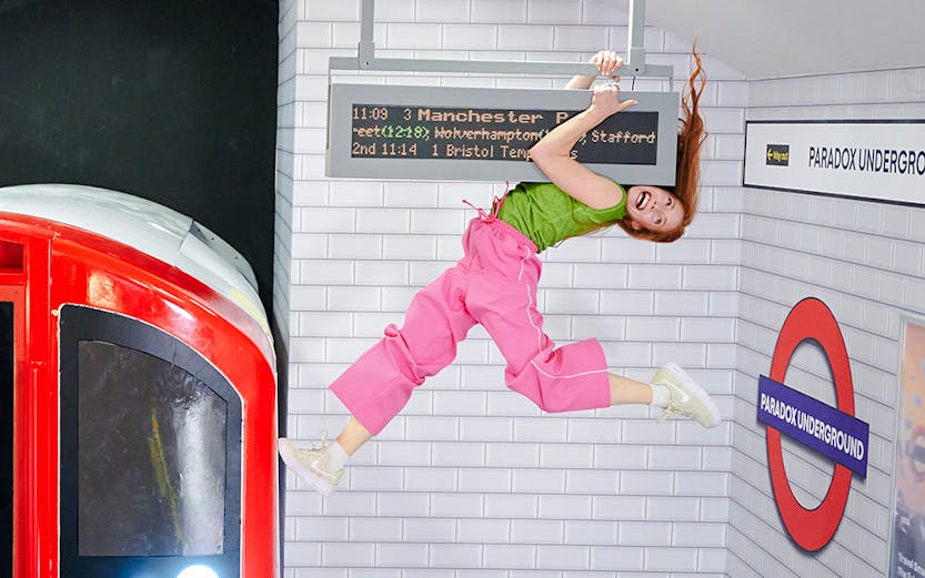 Guest hanging from a sign in a tilted subway illusion at Paradox Museum London.