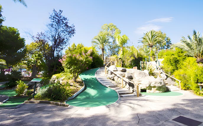 Mini-golf course with lush greenery and rock formations under a clear blue sky.
