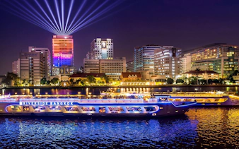 Riverside cruise ship on Chao Phraya River with Bangkok skyline illuminated at night.