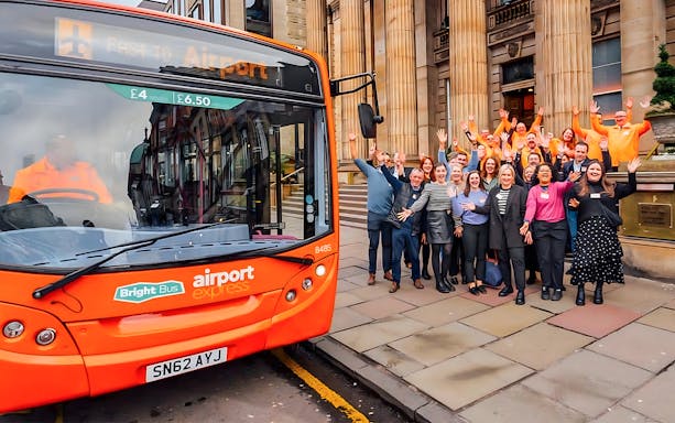 Bright Bus airport express in Edinburgh with a group of people waving outside a historic building.