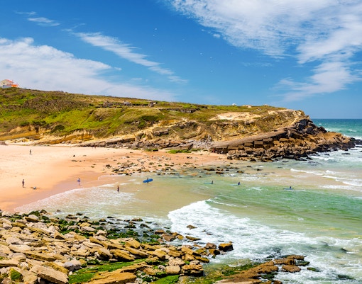 Beach near Pinhal da Nazare, Sintra, Portugal with surfers and rocky shoreline.