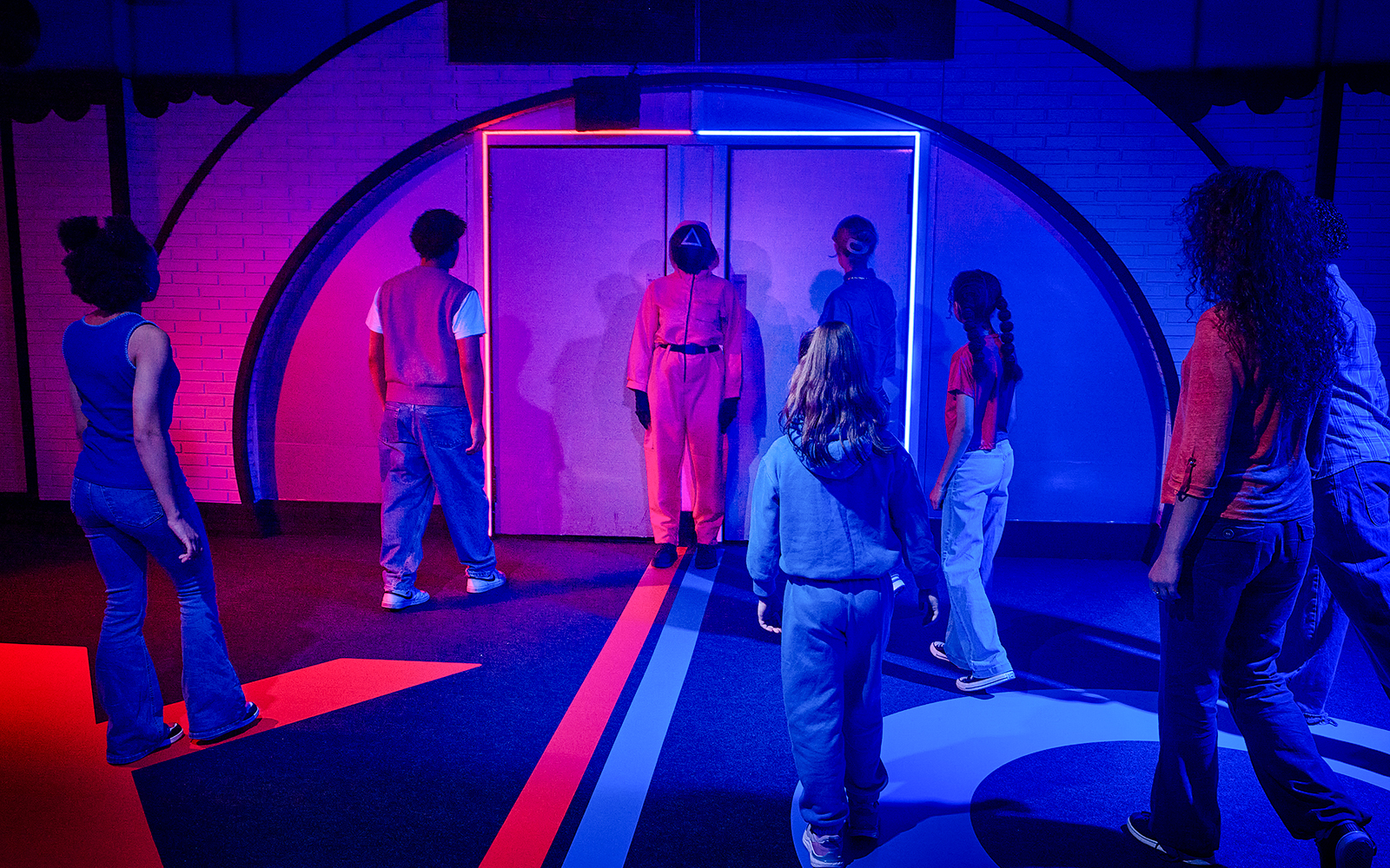 Participants in a Squid Game-themed experience in London facing a guard in a neon-lit room.