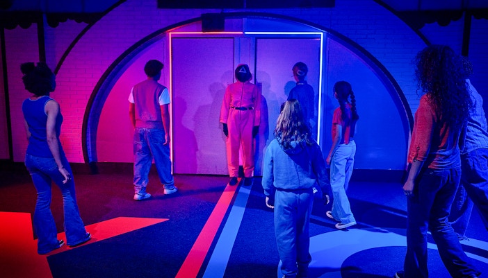 Participants in a Squid Game-themed experience in London facing a guard in a neon-lit room.