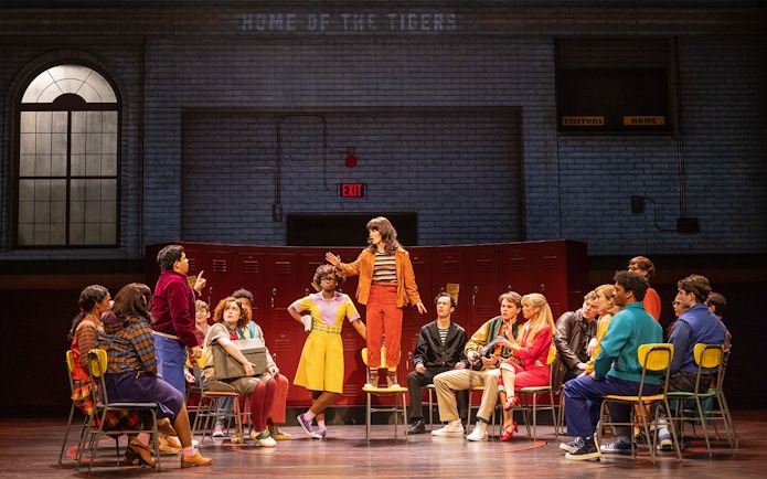 Group of people in a school gym setting, discussing around chairs, related to Stranger Things.