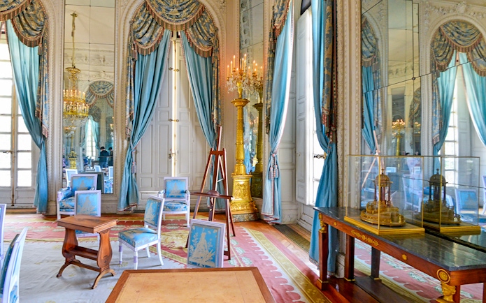 Grand Trianon room with ornate blue drapes, gilded candelabra, and elegant furniture.