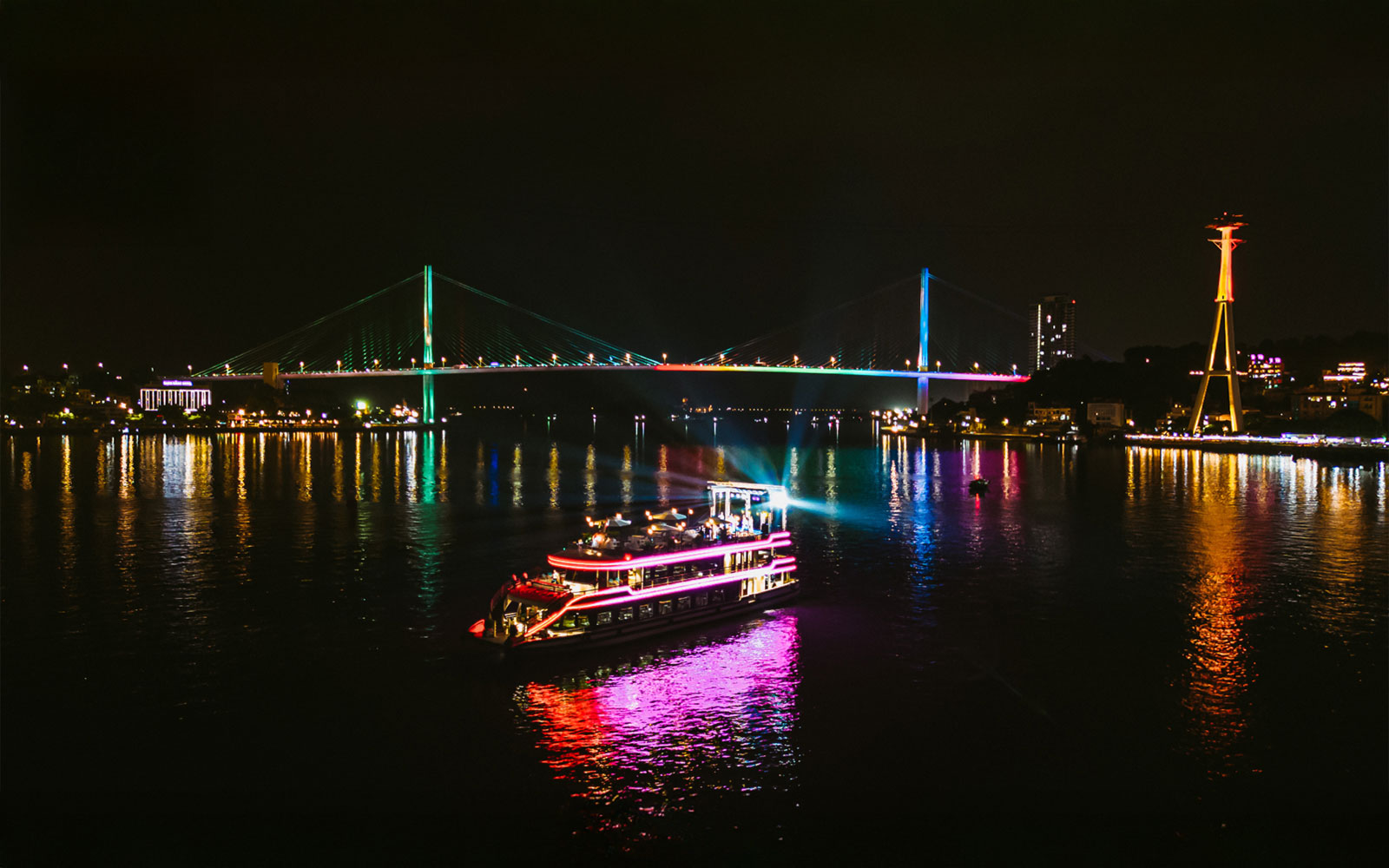 Cruise ship with colorful lights on Ha Long Bay at night, near illuminated bridge.