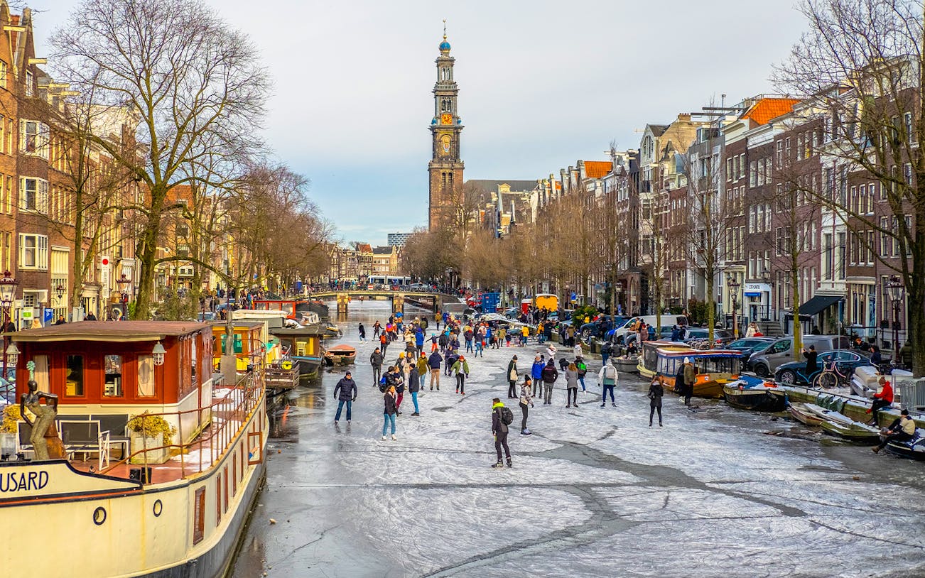 People ice skating on a frozen canal in Amsterdam with Westerkerk in the background.