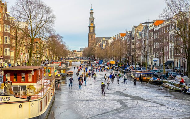 People ice skating on a frozen canal in Amsterdam with Westerkerk in the background.