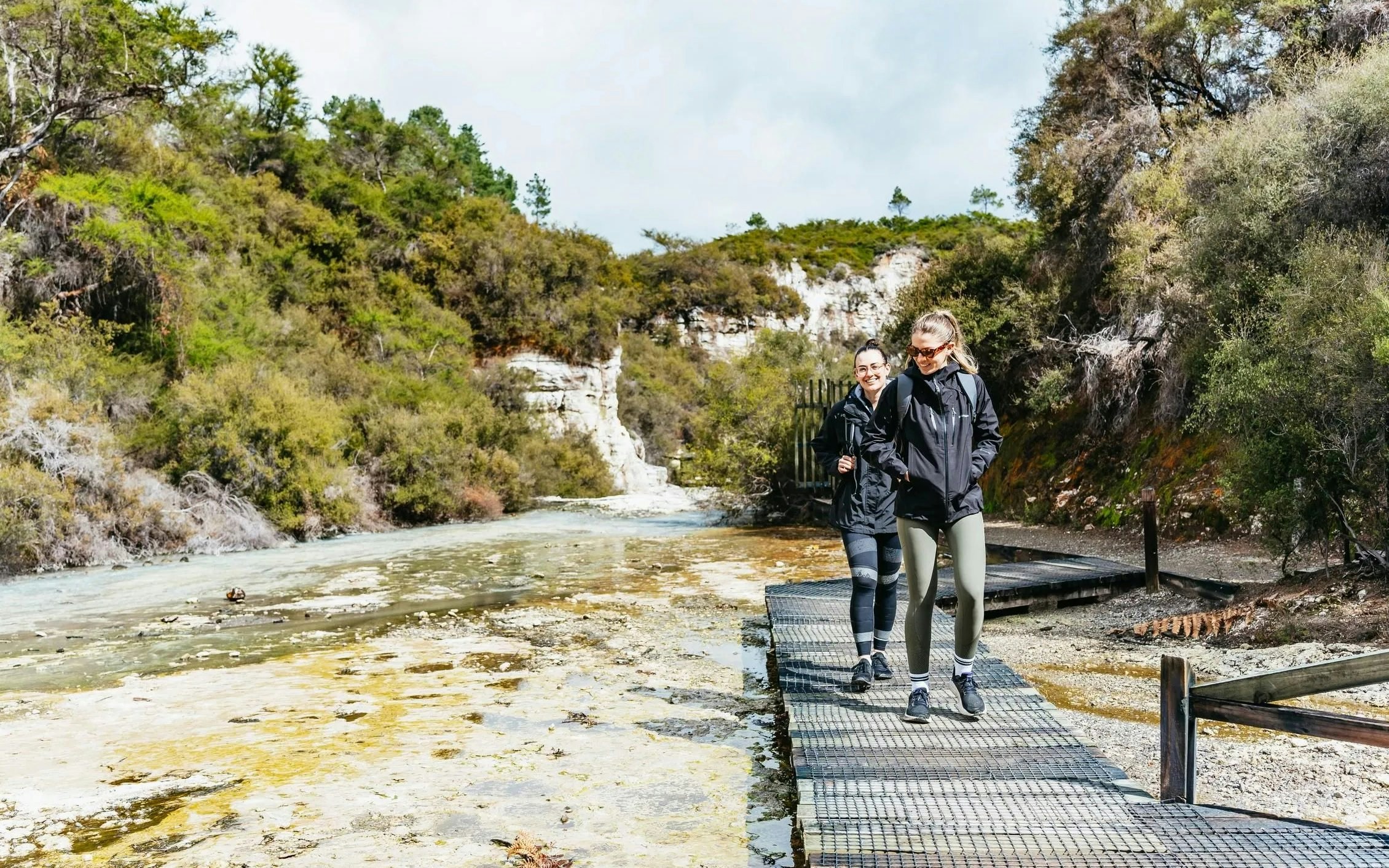 Visitors walking on a boardwalk at Wai-O-Tapu Thermal Wonderland, surrounded by geothermal landscape.