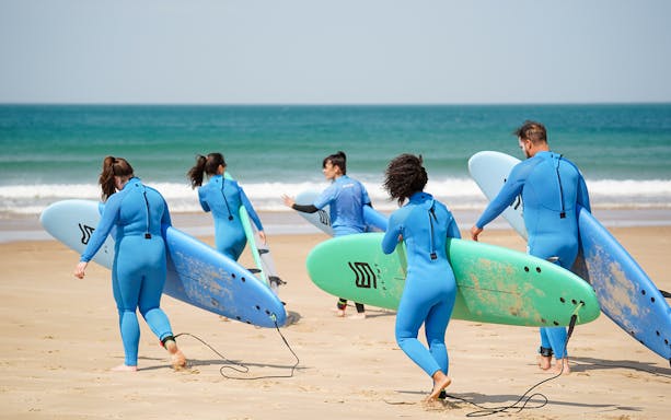 Surfers in wetsuits carrying boards on a Lisbon beach during a surf lesson.