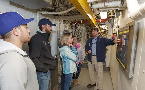 Guided tour group in engineering facility with guide explaining control panel.