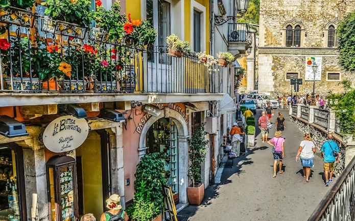 Narrow alleyway in Taormina with colorful flowers and people walking.