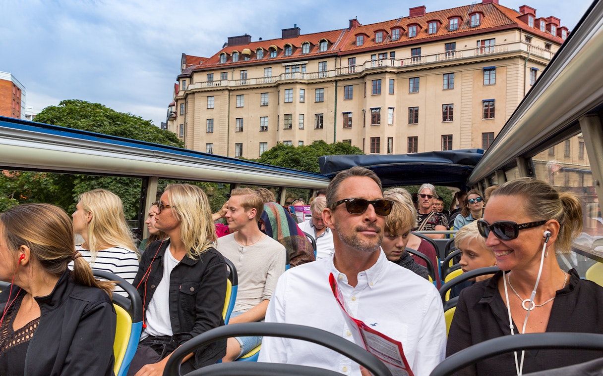 Open-top bus tour with passengers in Stockholm, historic building in background.