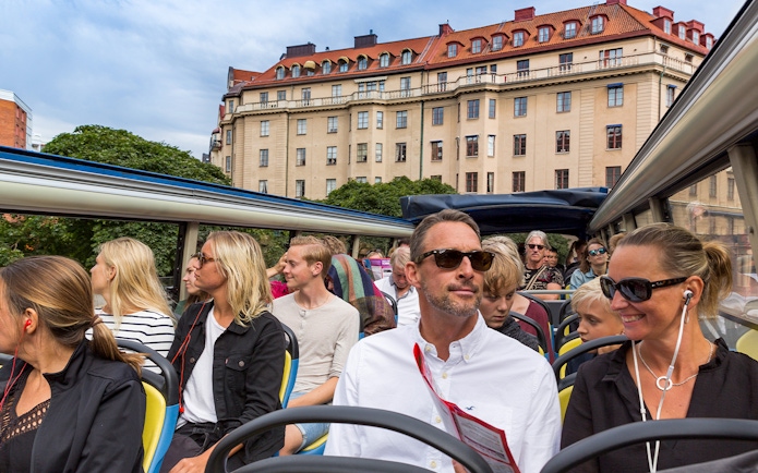 Open-top bus tour with passengers in Stockholm, historic building in background.