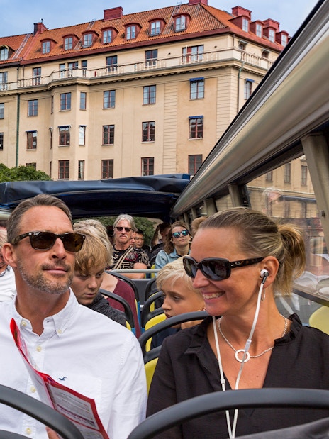 Open-top bus tour with passengers in Stockholm, historic building in background.