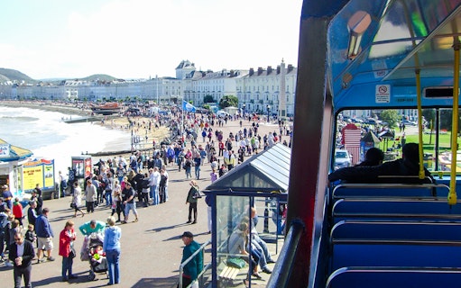 Tourists on an open-top bus in Llandudno, Wales, overlooking the promenade and beach.