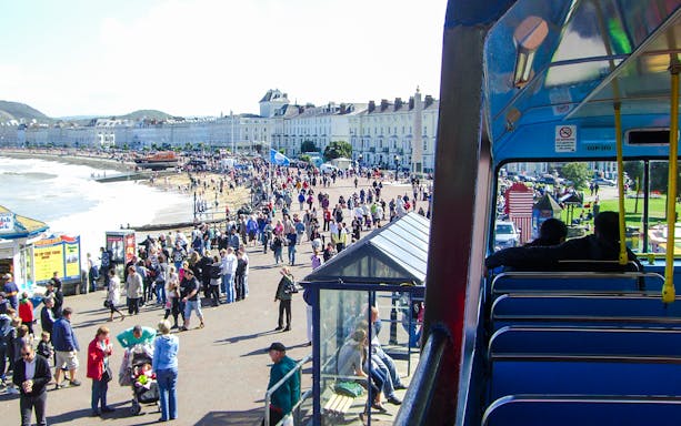 Tourists on an open-top bus in Llandudno, Wales, overlooking the promenade and beach.