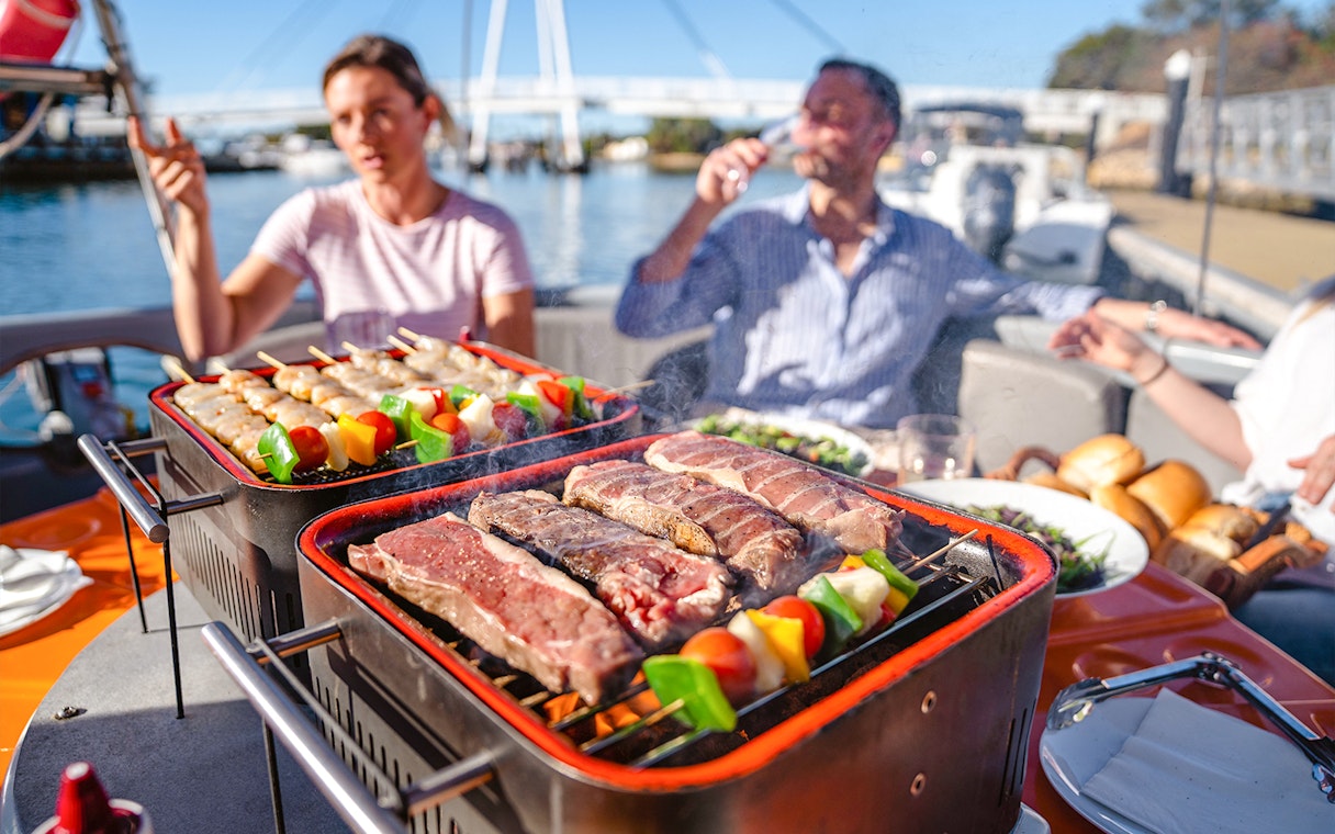 BBQ grill with skewers and steaks on a boat in Mandurah, people enjoying the self-drive tour.