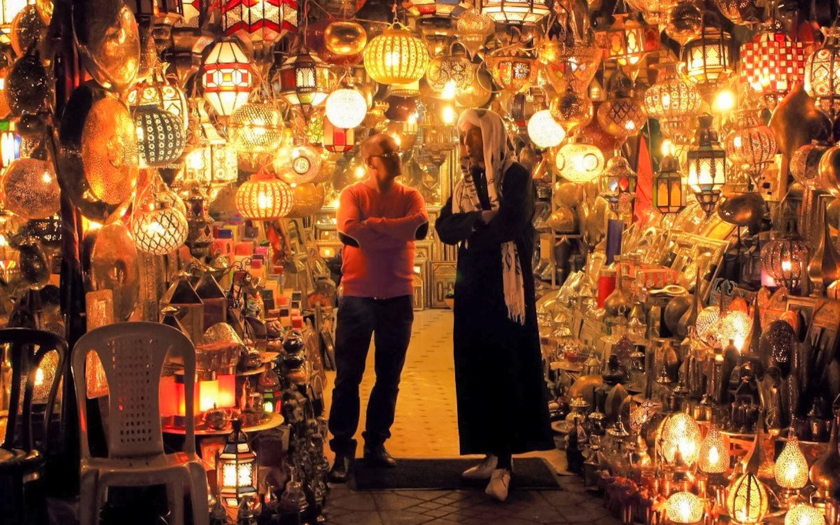 Moroccan market with colorful lanterns and two people conversing, related to vegetable tagine tour.