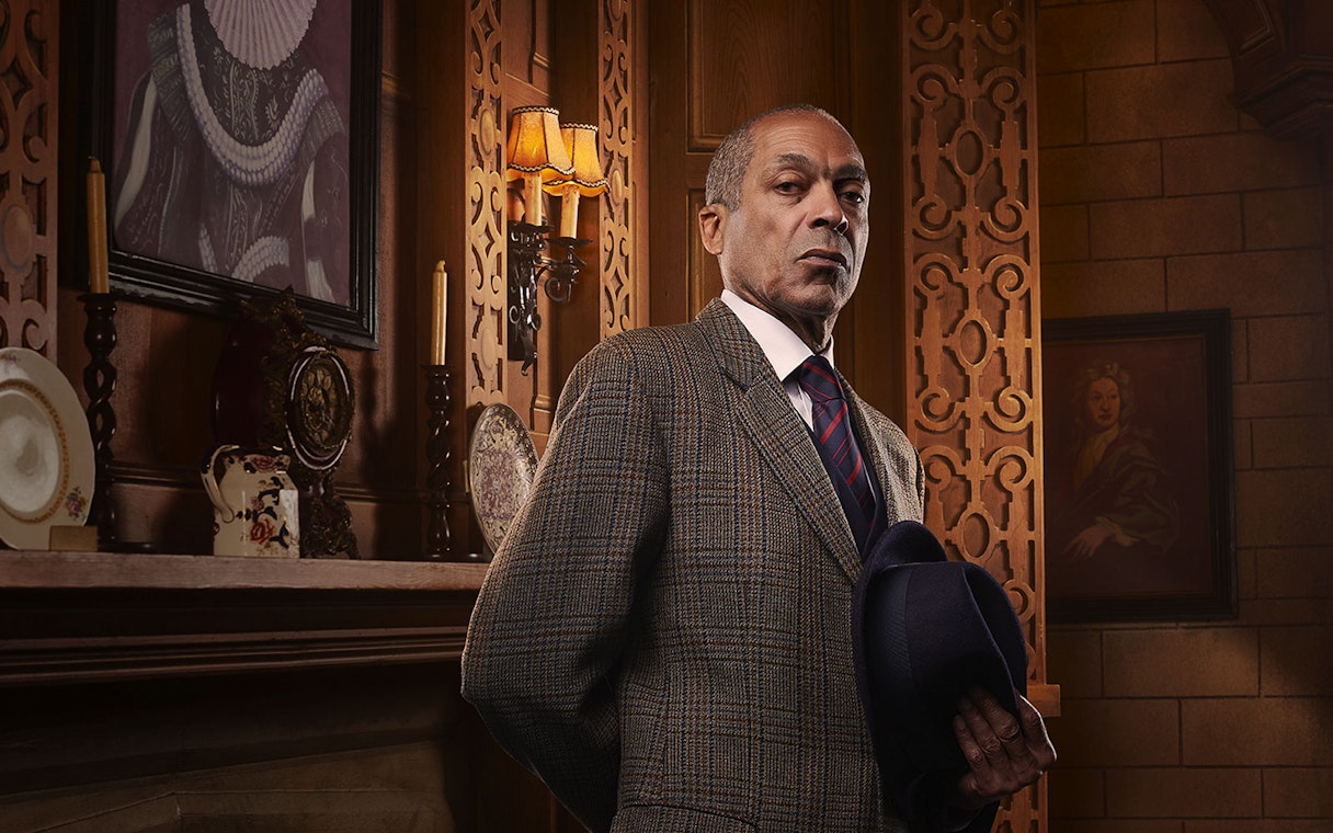 Man in a suit holding a hat, standing in an ornate room with vintage decor, related to The Mousetrap.
