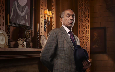 Man in a suit holding a hat, standing in an ornate room with vintage decor, related to The Mousetrap.