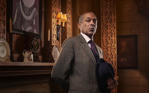 Man in a suit holding a hat, standing in an ornate room with vintage decor, related to The Mousetrap.