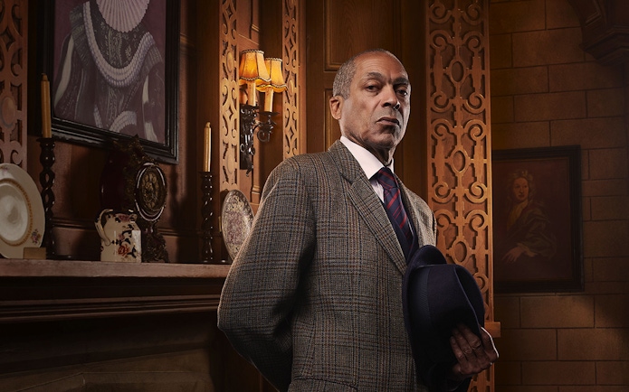 Man in a suit holding a hat, standing in an ornate room with vintage decor, related to The Mousetrap.