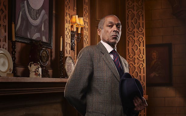 Man in a suit holding a hat, standing in an ornate room with vintage decor, related to The Mousetrap.