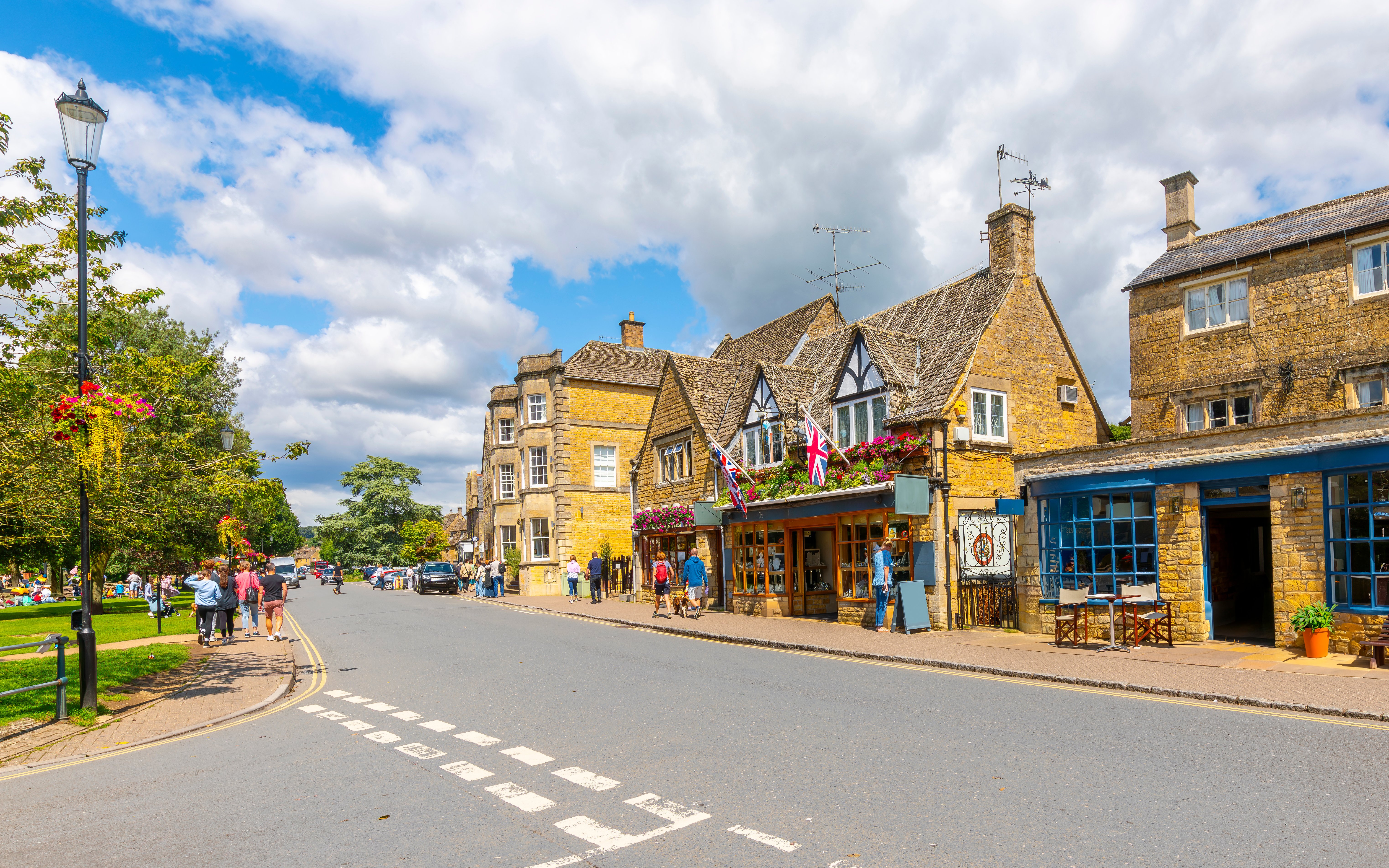 Cotswold village street with stone buildings and people walking.
