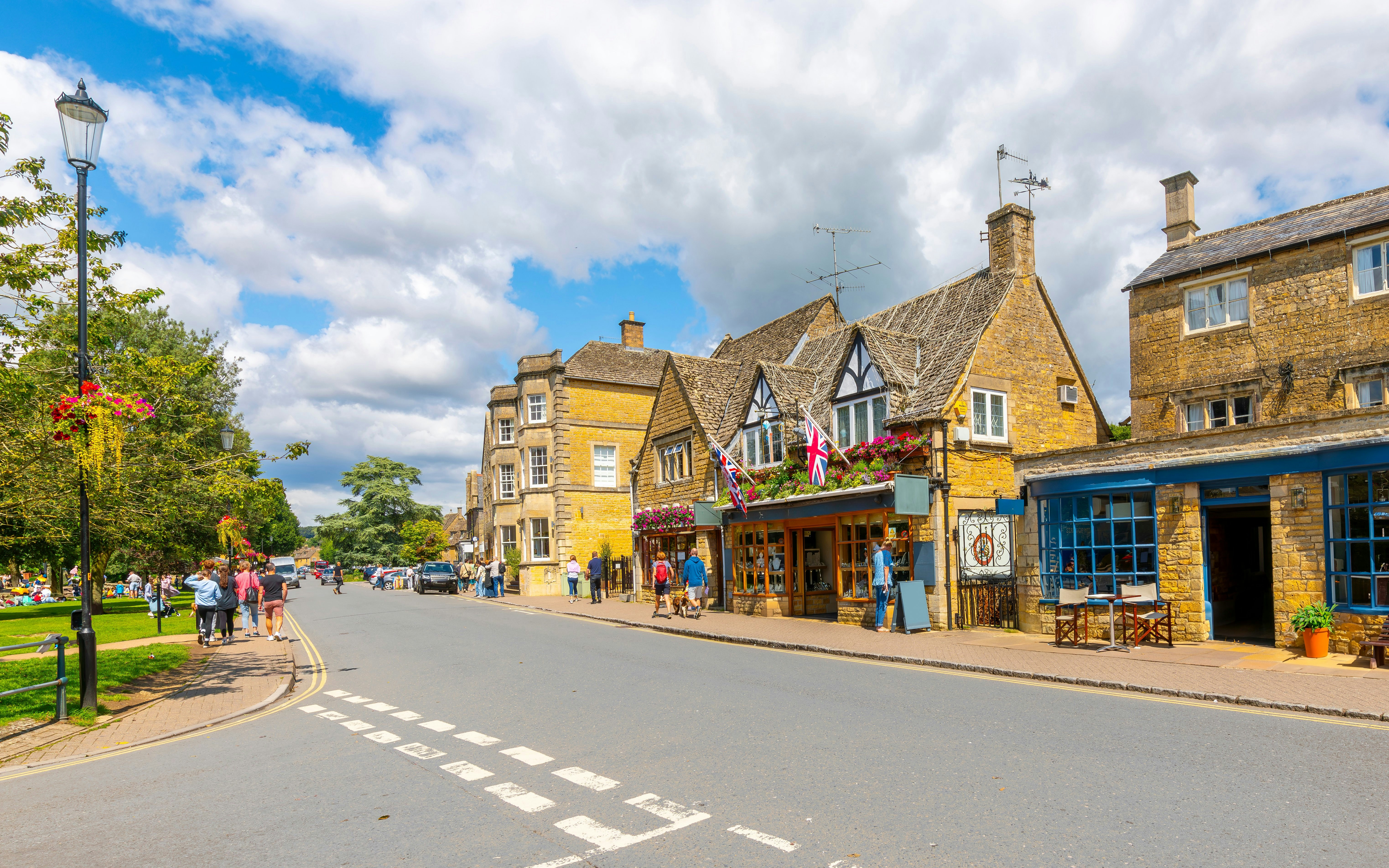 Cotswold village street with stone buildings and people walking.