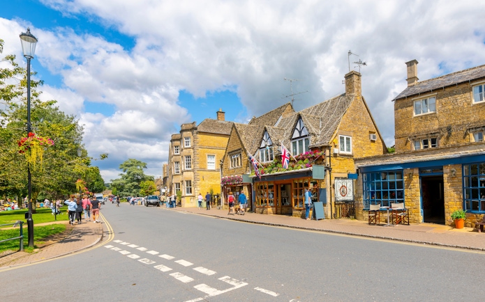 Cotswold village street with stone buildings and people walking.