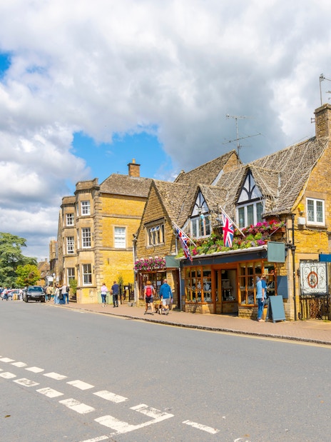 Cotswold village street with stone buildings and people walking.
