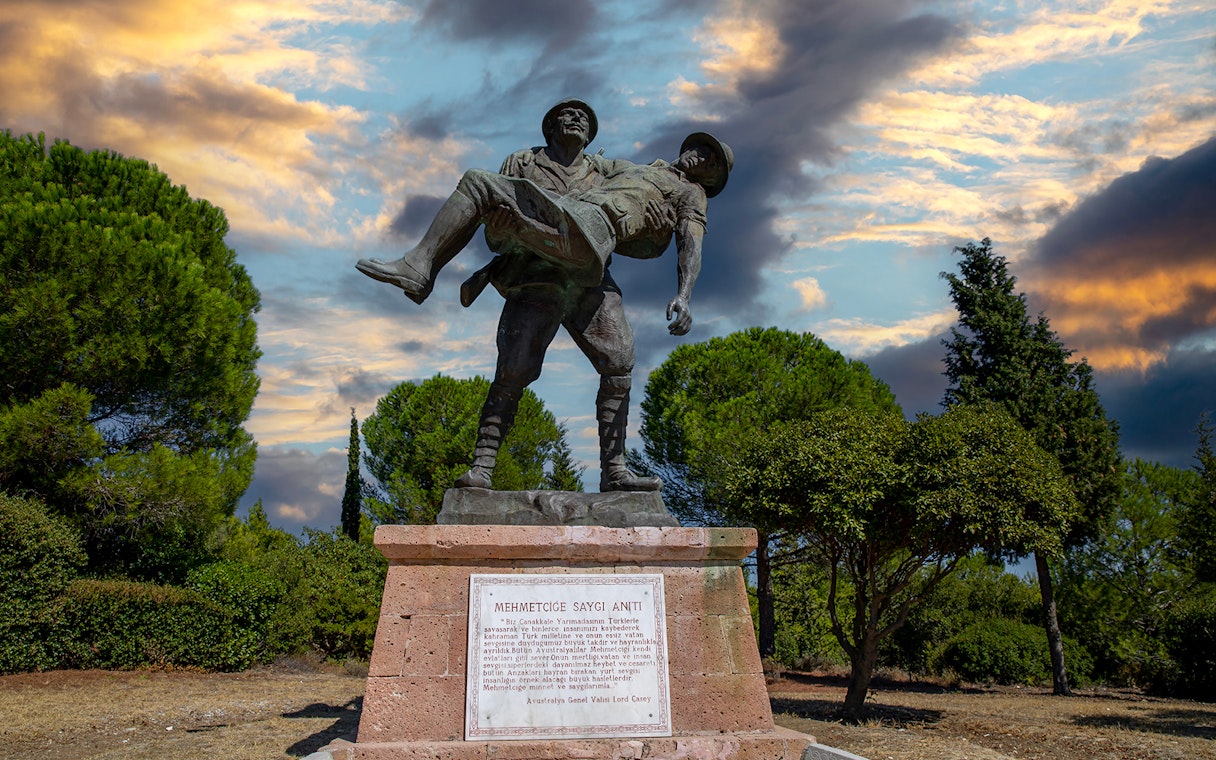 Gallipoli statue of soldier carrying comrade, surrounded by trees, under a dramatic sky.