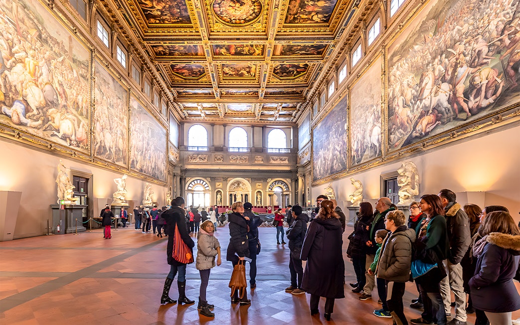 Visitors inside the ornate hall of Palazzo Vecchio, Florence, viewing large frescoes and sculptures.