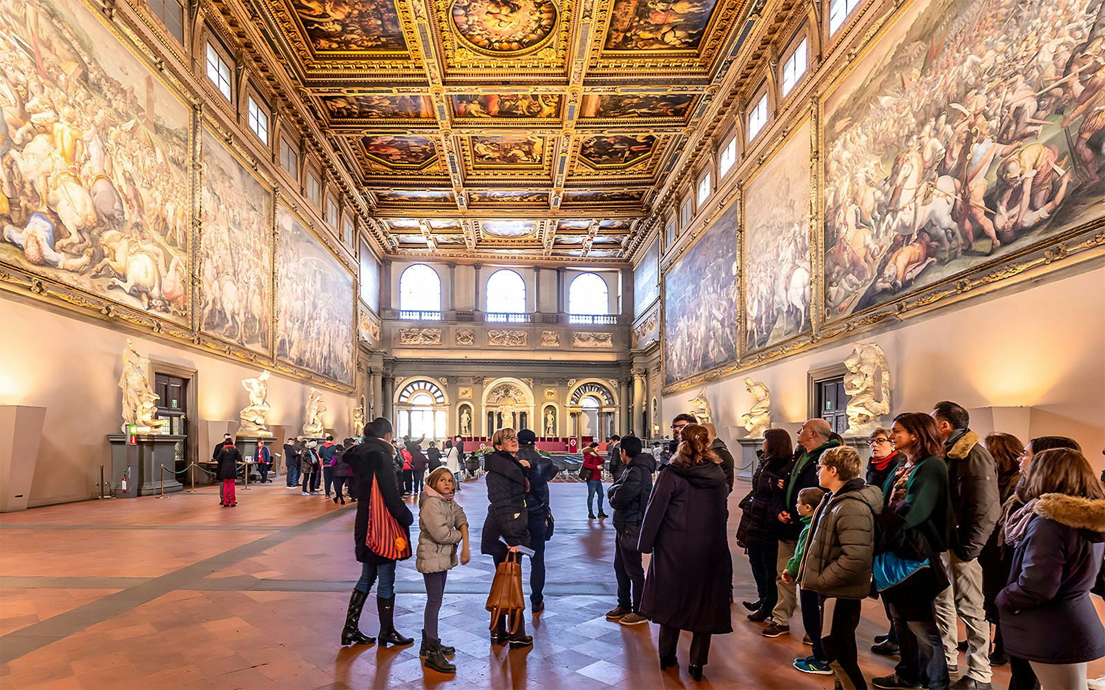 Visitors inside the ornate hall of Palazzo Vecchio, Florence, viewing large frescoes and sculptures.