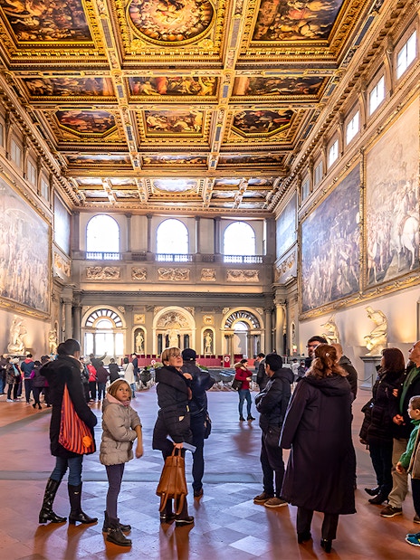 Visitors inside the ornate hall of Palazzo Vecchio, Florence, viewing large frescoes and sculptures.
