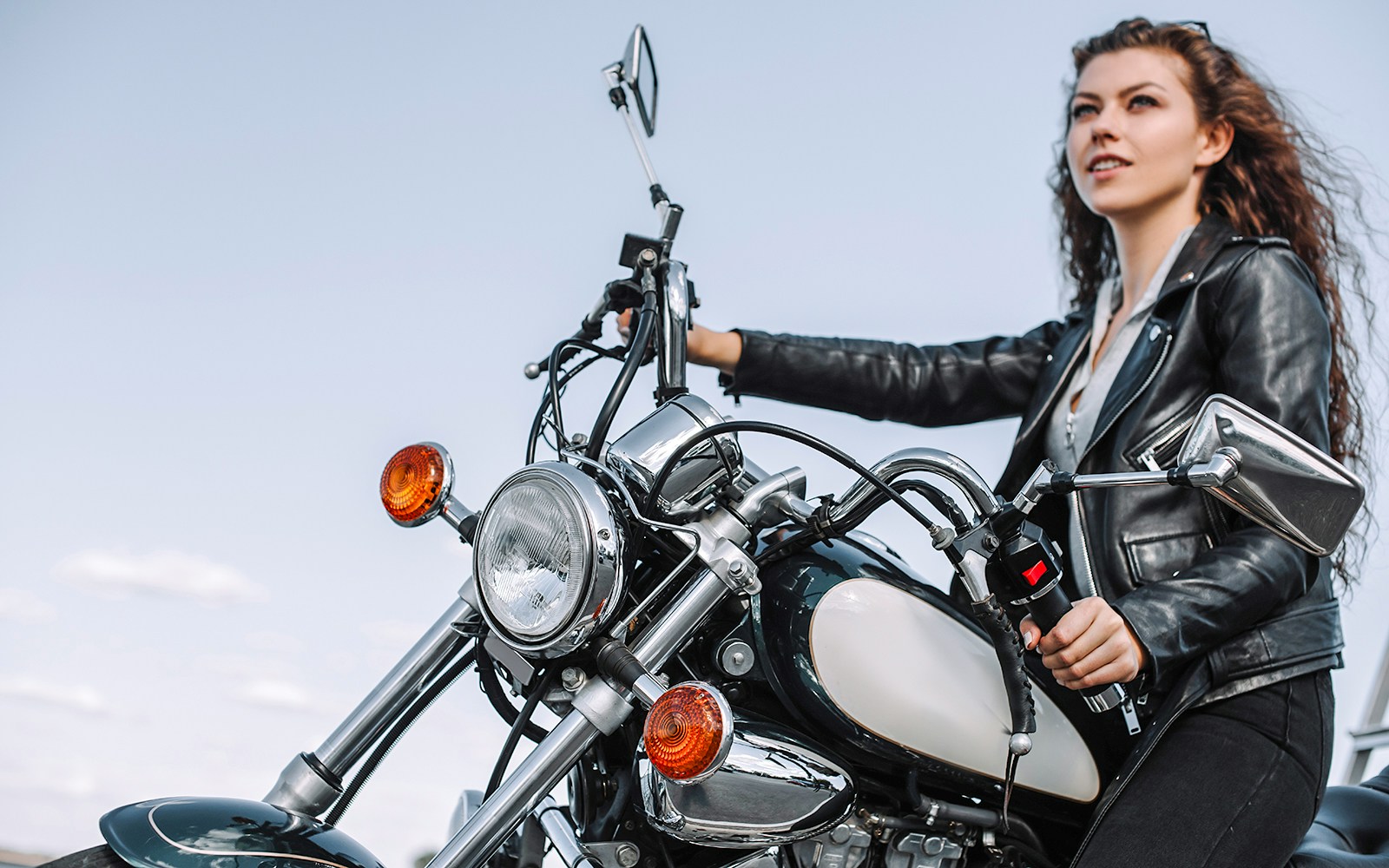 Girl riding a motorbike in a leather jacket against a clear sky.