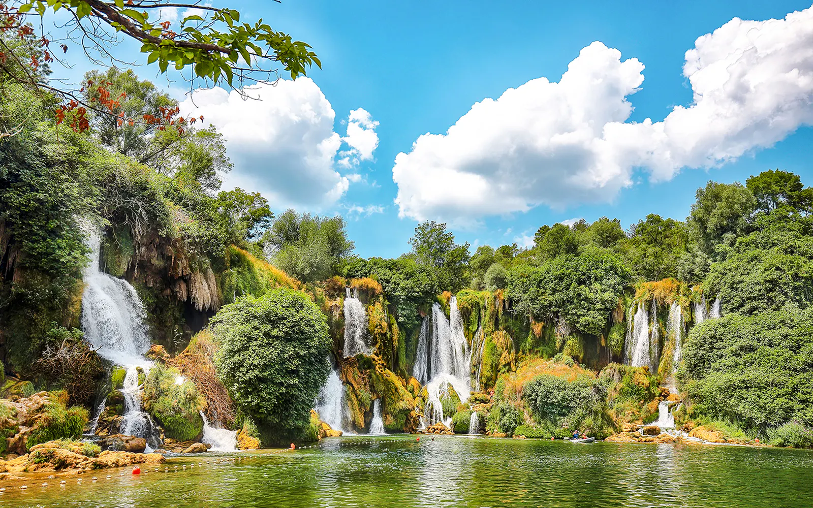 Kravice Waterfall cascading into a lush green pool surrounded by dense foliage.