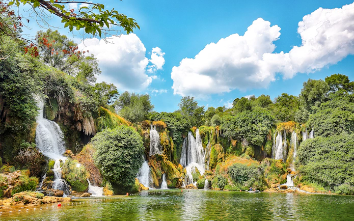 Kravice Waterfall cascading into a lush green pool surrounded by dense foliage.