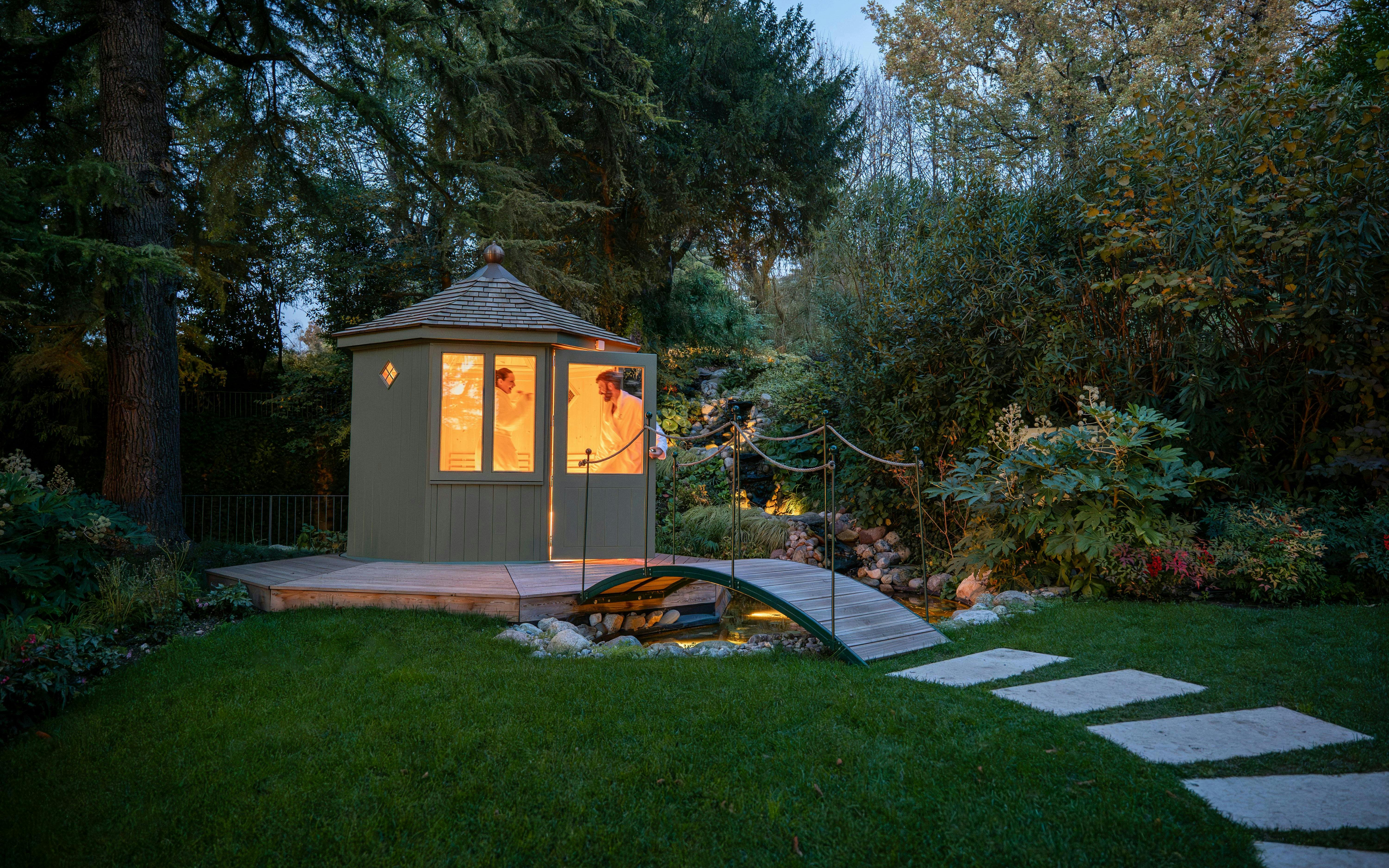 Exteriors of QC Terme Garda with illuminated cabin and garden bridge at dusk.