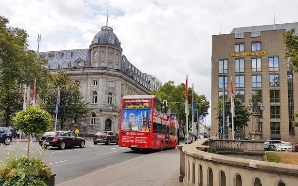 Red double-decker bus on a city tour in Düsseldorf, passing historic buildings.