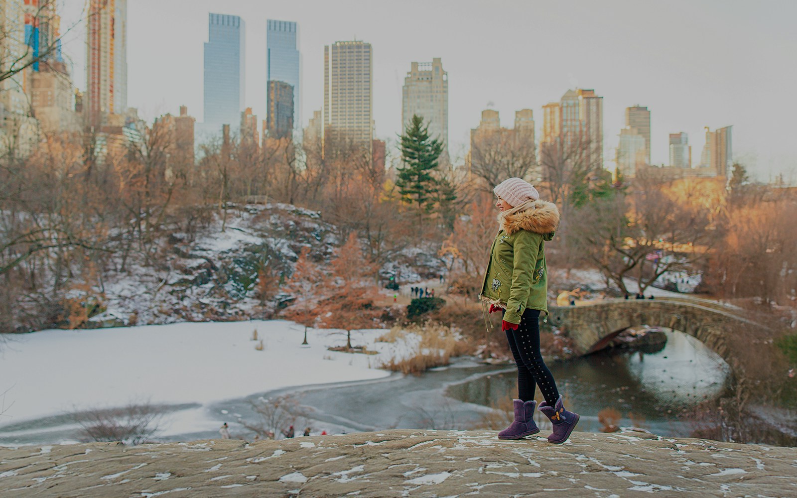 Girl standing in Central Park, New York City, with a view of the skyline and a stone bridge.